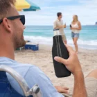 Man on beach chair holds an Elmer Bottle Buddies; two talk by the ocean in the background.