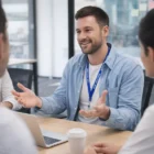 Man smiling, talking with colleagues at table with laptop, coffee, and 10Mm Silicon Lanyards.