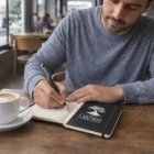 Man writing in a Zevar Notebooks With Pens labeled Lake Tree Landscaping at a cafe with coffee.