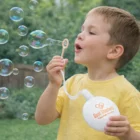 Young boy in yellow shirt blowing bubbles outdoors with Zorbs Bubble Kits wand and container.