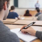 Student taking notes with Elixion Pens in class, other students and teacher in background.