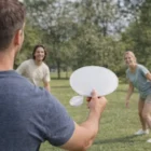 Three play paddleball as Avant Fans With Pouches rest nearby on the sunny park grass.
