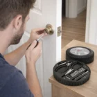 Person fixes door lock with screwdriver; Tyre Shaped Tool Kit is on the table nearby.