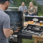 Man grills burgers with Premium Promo Bbq Sets; two people relax in the background.