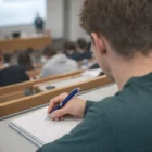 Student taking notes with Corporate Printed Cupid Pens in a lecture hall with teacher and classmates.