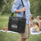 Man carries Large Icy Cooler Bag in park; picnic and basket in background.