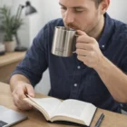 Man sipping from a Stainless Steel Logo Engraved Mug while reading at a desk with laptop and notebook.