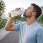 Man drinks from an Alaska Protein Shaker outdoors on a scenic Alaskan pathway.