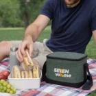 Man on picnic blanket grabs sandwich near fruit, water, and Travel Pack Compact Chiller Bag.