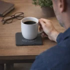 Someone with a coffee mug on Nodex Coasters at a wooden table with glasses and a book.