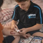 Boy applies Temporary Tattoos Classic 51Mm X 76Mm to friend’s arm while on the bed.