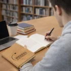 Writing in a Medium Cork Cover Notebook at a library table with books, laptop, and coffee.