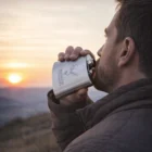Man drinks from a Jefferson Custom Engraved Hip Flask at sunset outdoors in a brown jacket.