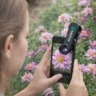 Person uses a 3-In-1 Smartphone Lens Kit to photograph pink flowers in a garden.