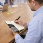 Man writing in a Denver Branded Notebook at a meeting table with laptops and sticky notes.