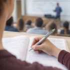 Student takes notes with Best Value Logo Event Pens in a lecture hall as the teacher speaks.