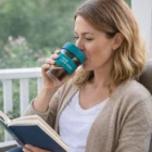 Woman reads a book, sipping from Clear Reusable Coffee Cup with blue lid and logo.