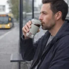 Man at bus stop drinks coffee from 350Ml Apollo Custom Glass Reusable Cup, bus in background.