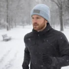 Man jogging in a snowy park, wearing a black jacket and Extra Warm Logo Patch Beanies.
