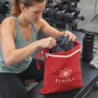 At the gym, a woman puts clothes into a red Eureka Wet Bags with dumbbells beside her.