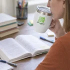 Woman in orange sweater drinks from a white Zesty Vacuum Cup With Handle while studying.