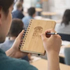 Person writing in an Edmon Bamboo Notebook labeled SKY RESORT THAILAND HEALTH in class.