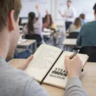 Student writing in a Trainor Paper Finish Notebook labeled STEAM EDUCATION in class with peers.