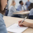 Person writes notes with a Zyd Ball Pen; others sit at tables in the background.