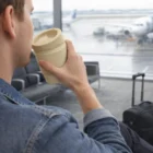 Man with 340Ml Bamboo Blend Reusable Cup at airport gate, suitcase beside him, planes outside.