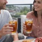 Two smiling people hold Ofolia Glass Sets 410Ml of iced tea by fruit and pastries outdoors.