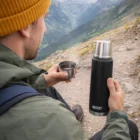 Person in outdoor gear holds a Swiss Peak Elite Copper Vacuum Flask, enjoying a mountain view.