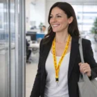 Smiling woman in business attire with a yellow Zairi 3D Logo Lanyard, carrying a gray tote bag.