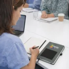 At a meeting, a woman writes in a Heather Notebook And Pen Gift Set open on the table.