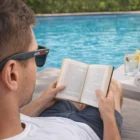 Man in Lagoon Premium Sunglasses Carbon Fibre reads by the pool, drink on table nearby.