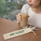 Woman sipping iced coffee with an Etello Steel Straw Set; farmers market pouch on table.
