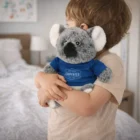 Young child hugs a Cuddly Koala Plush Toy while wearing a blue Carpenter shirt in the bedroom.