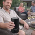 Smiling man holds black tumbler; friends with wine glasses and a Lyon Vacuum Wine Cooler.