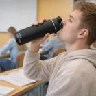 Young man in hoodie drinks from a Swiss Peak Vacuum Bottle in a classroom with other students.