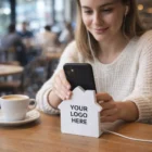 Woman charging phone with Alicante Wireless Charger, house-shaped and labeled YOUR LOGO HERE.