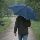 Person walks in rain with a large blue Blunt Coupe Umbrella on a park path.