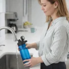 Woman fills a blue Promotional Camelbak 750Ml Mag Bottle at a kitchen sink with modern faucet.