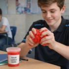 Smiling teen squeezes red Promotional Stress Slimes; "Your Logo Here" on container on desk.