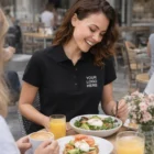 Smiling woman in a Sols Passion Womens Polo eating salad at an outdoor cafe, YOUR LOGO HERE.