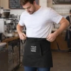 Man tying a Raya Full Length Waist Apron in a cozy coffee shop.