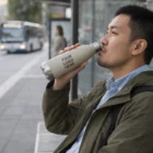 Man at bus stop drinks from Rushmoor Natura Bottle featuring Your Logo Here label.