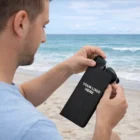 Man on beach holds Deane Sunglass Pouches with custom logo, ocean in background.