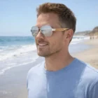 Man wearing Kent Mirror Lens Sunglasses Bamboo, smiling on a sandy beach with ocean behind.
