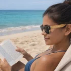 Woman in Joaquin Sunglasses Bamboo reading a book on a sandy beach by the ocean.