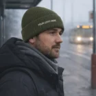 Man in winter coat and Wade Knitted Beanies with YOUR LOGO HERE, standing at a bus stop.
