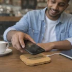 Man charging phone on Tasman Bamboo Wireless Chargers Square at cafe with coffee, notebook nearby.